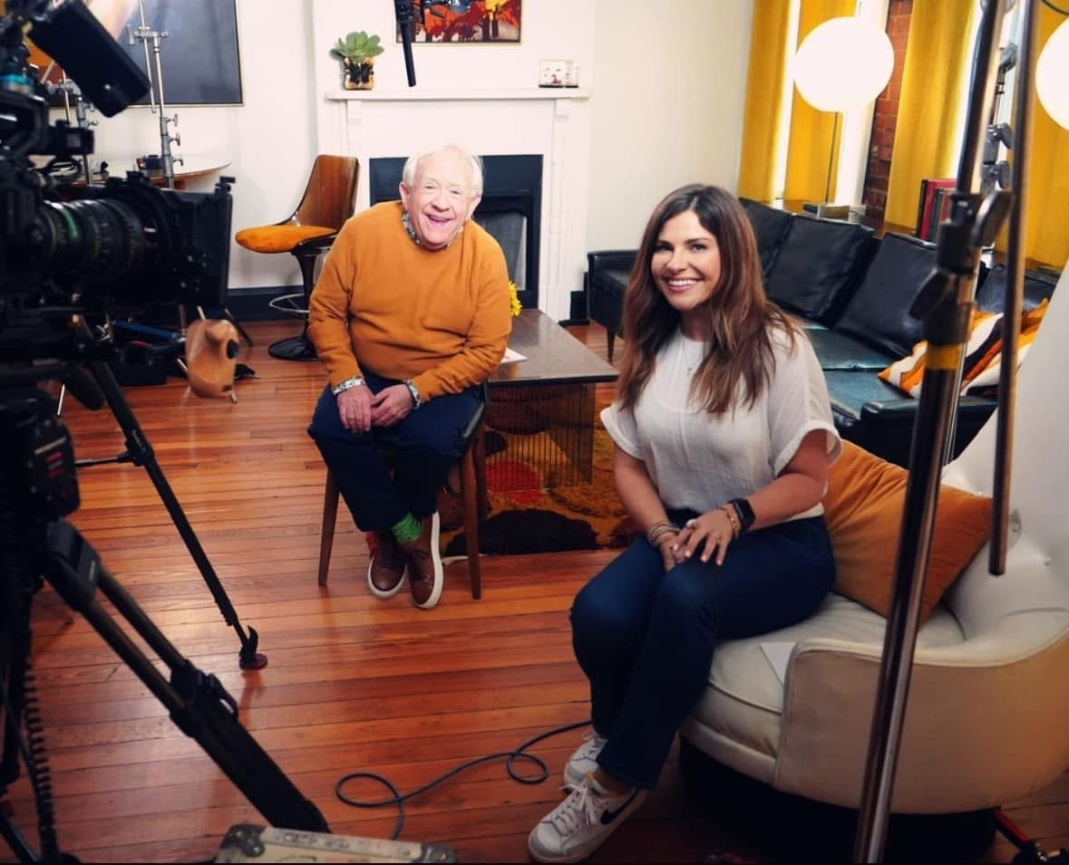 Two people seated in a studio with cameras, lighting, and a wooden floor during a filmed interview