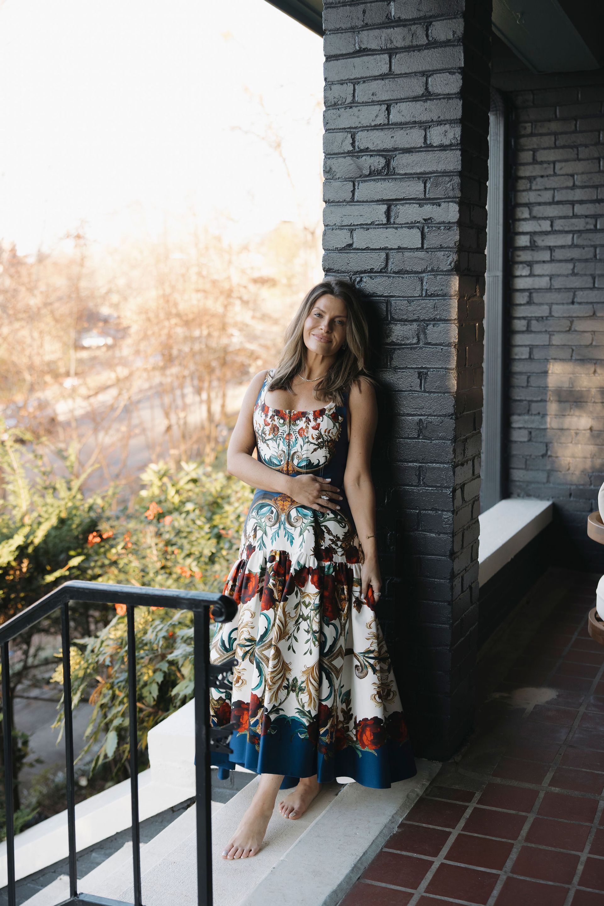 Woman in a floral dress leaning by a brick wall on a balcony, with autumn trees in the background.