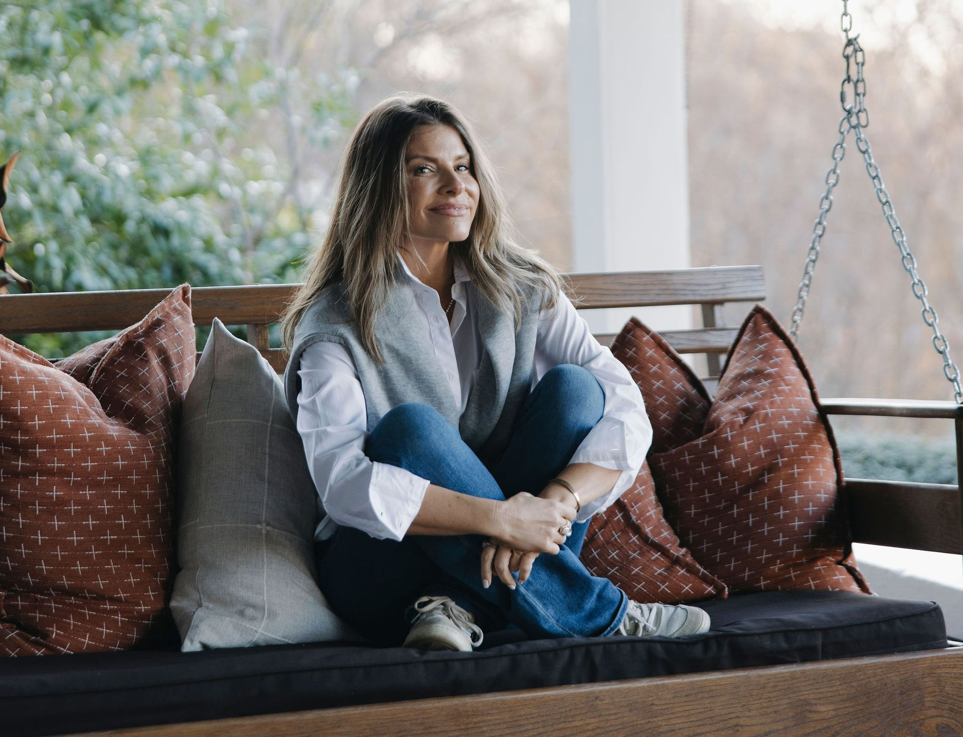 Woman sitting cross-legged on a cushioned porch daybed with patterned pillows, looking out a window.