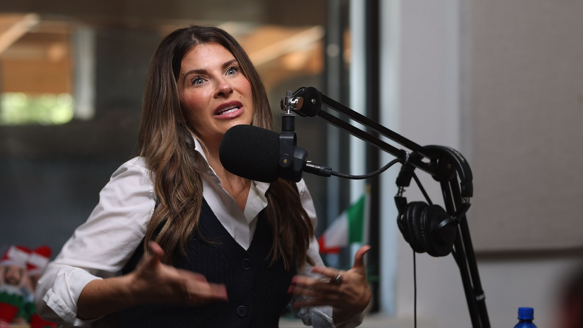 Woman speaking into a podcast microphone in a studio, gesturing with her hands.