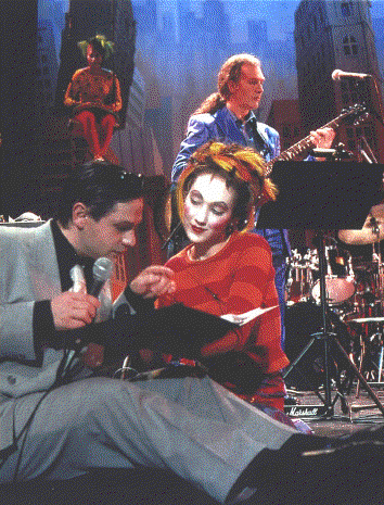 Band performing onstage; woman in red, man in suit looking at sheet music. Guitarist and another figure in background.