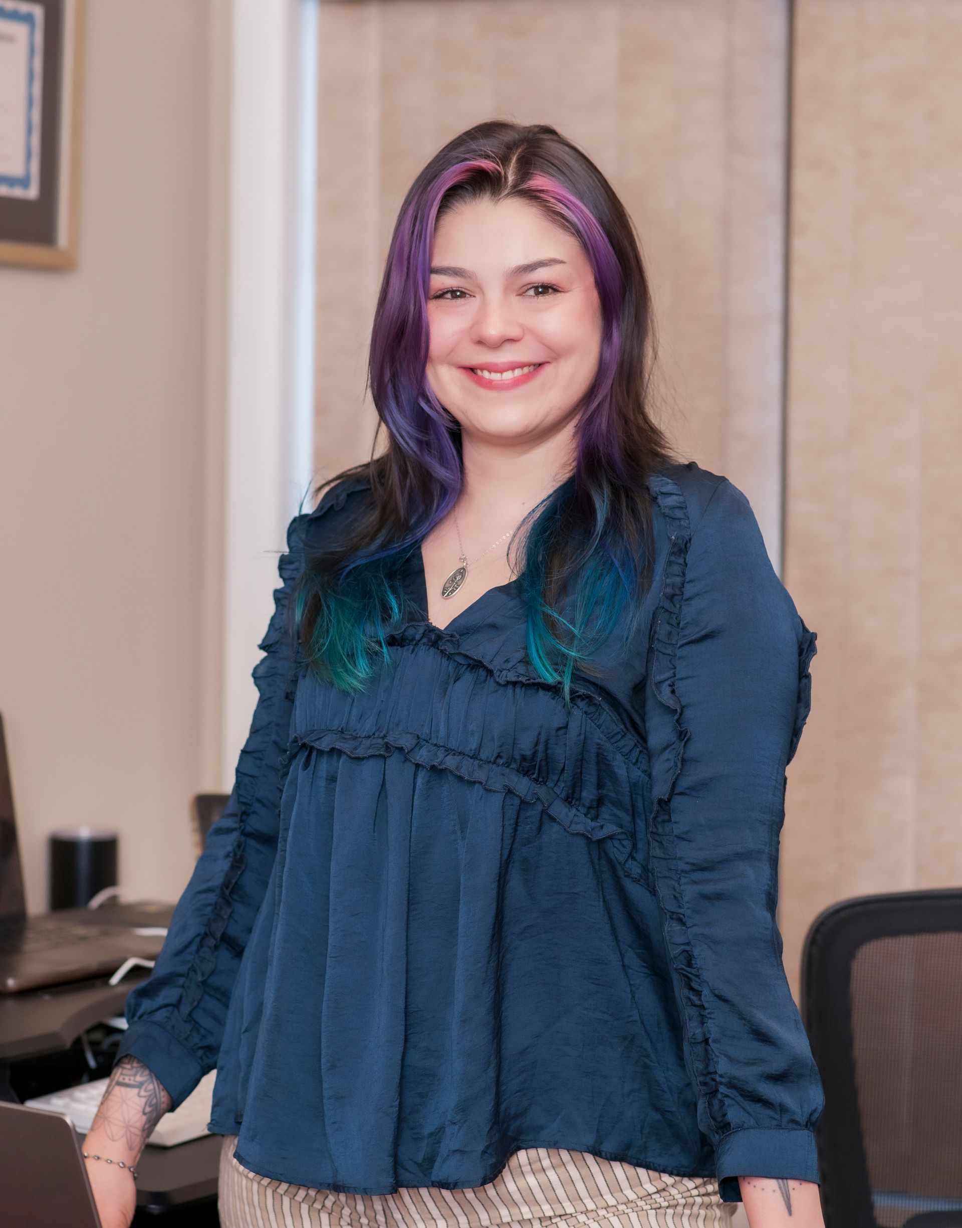 Woman with purple-streaked hair smiles, wearing a blue ruffled top, standing near a desk in an office.
