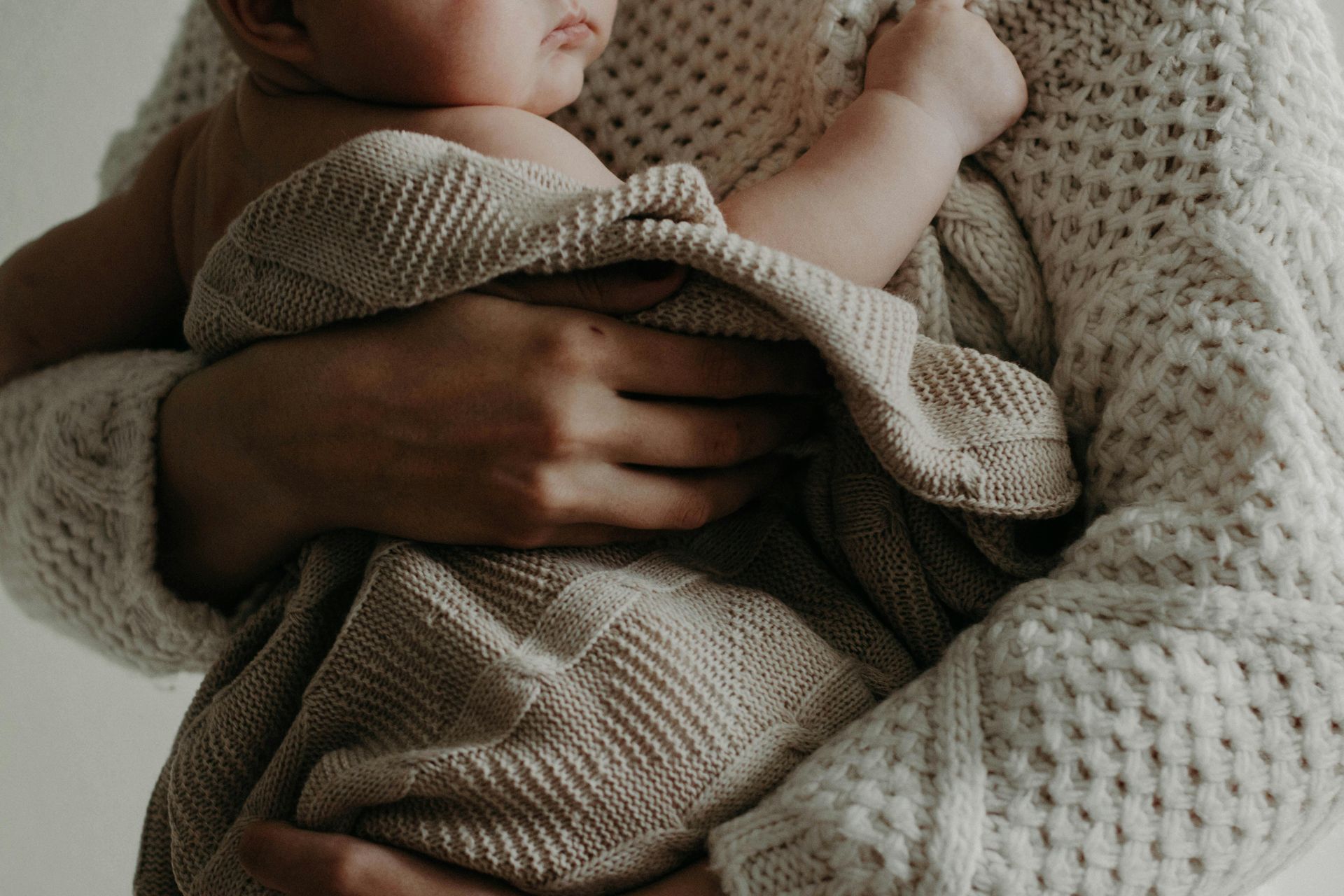 A person holding a swaddled baby; both wearing light-colored knitwear.