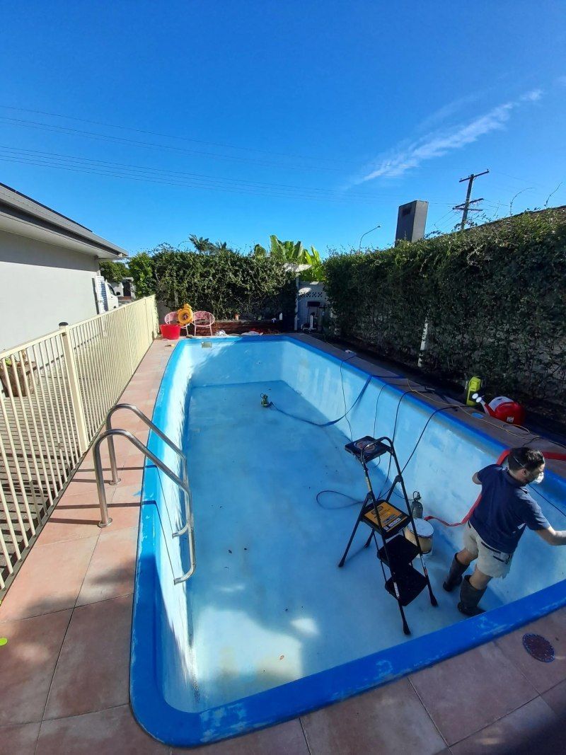 Worker cleaning an empty backyard pool with a hose and tools on a sunny day