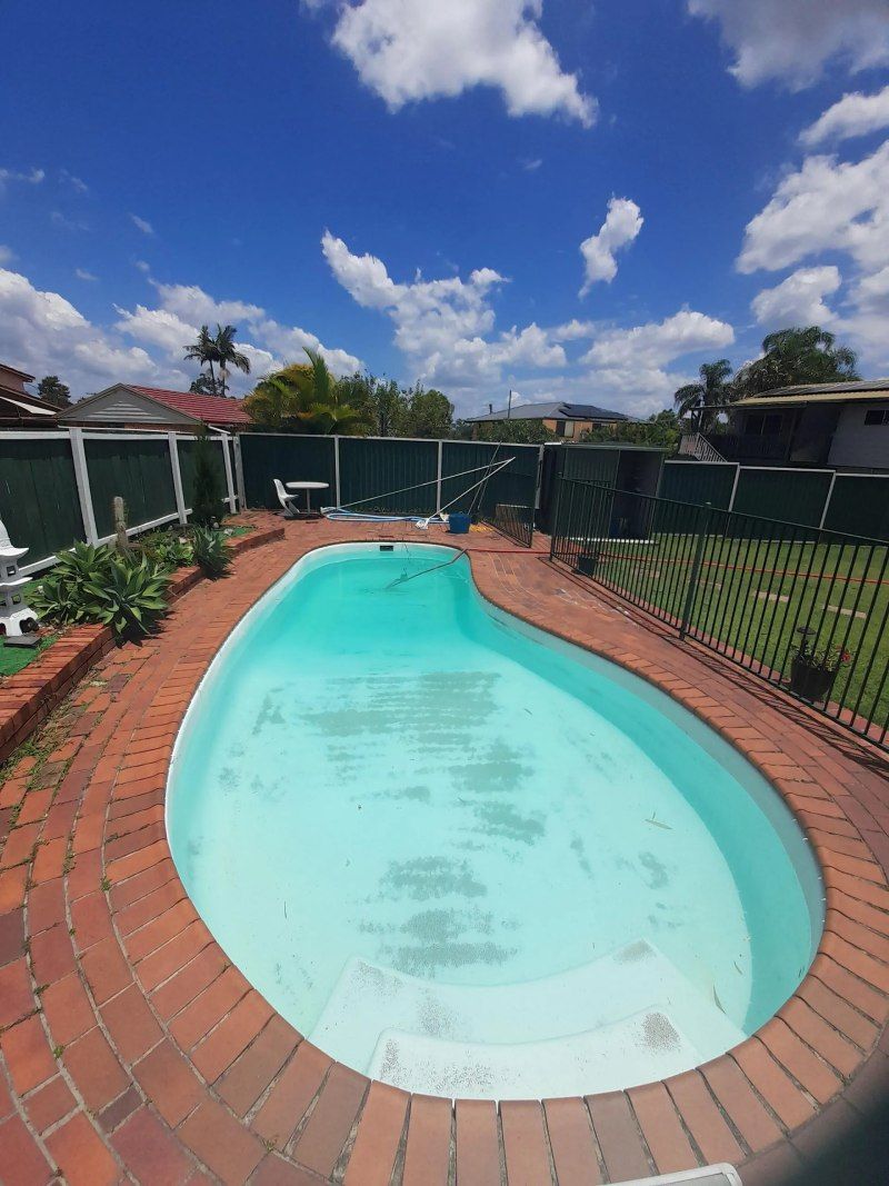 Backyard swimming pool with red brick deck, green fencing, and a bright blue sky with clouds.