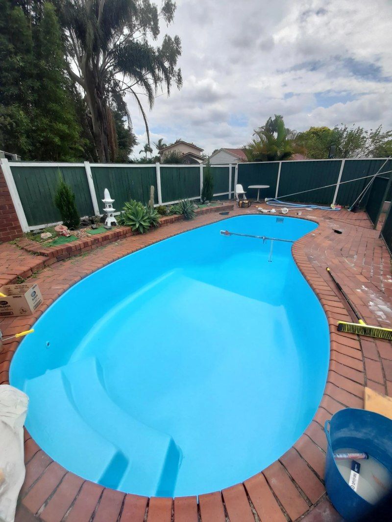 Backyard swimming pool with blue water, brick patio, and green fence on a cloudy day