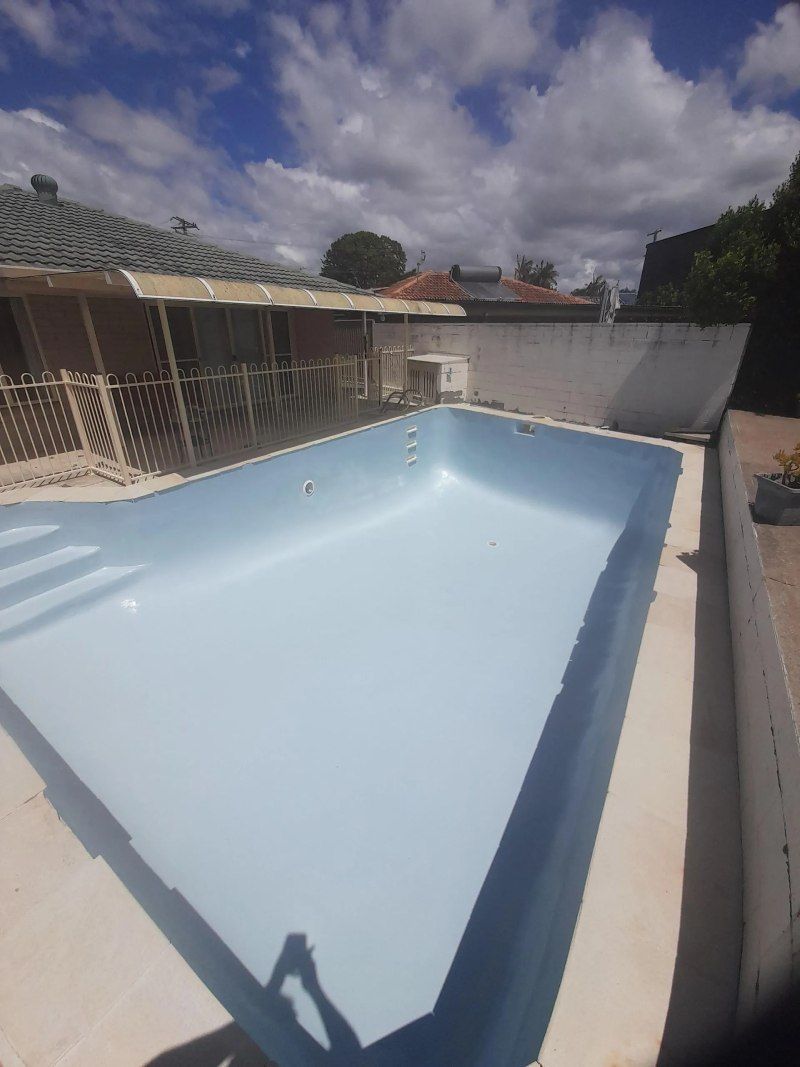 Empty light-blue swimming pool in a sunny backyard with a white wall and covered patio.