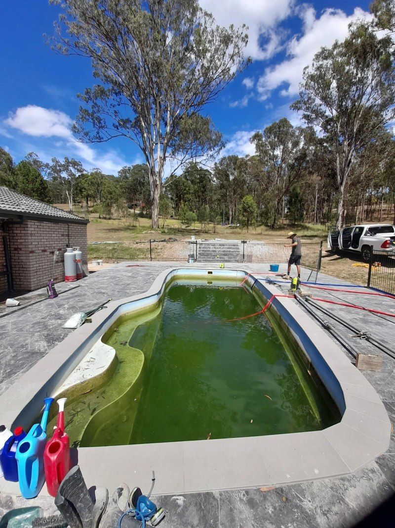 Small outdoor swimming pool under construction, with green water, paving stones, and trees in the background.