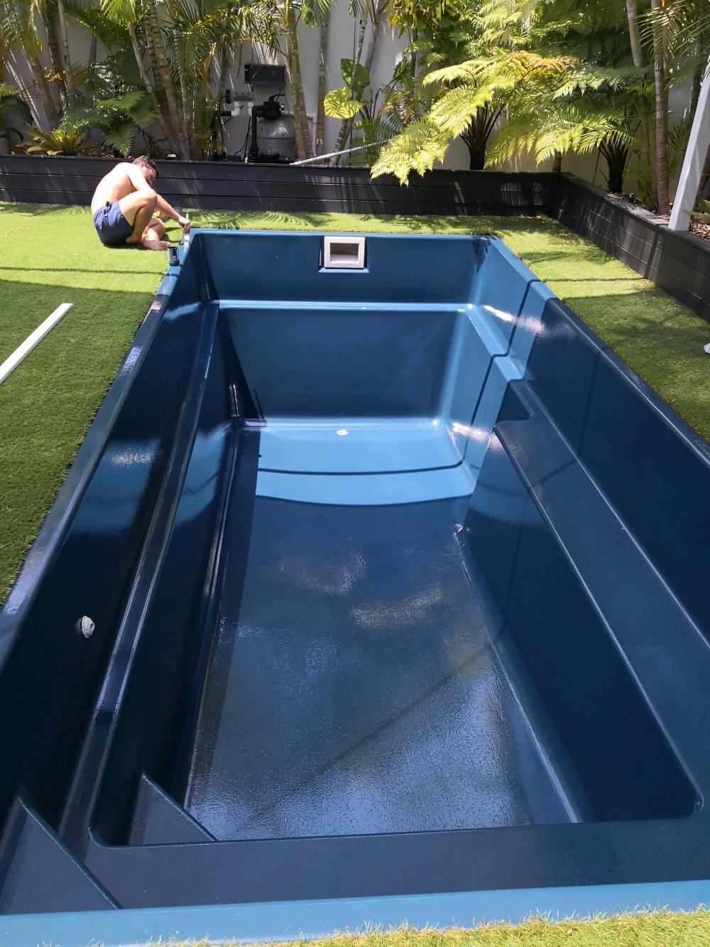 Blue swimming pool under construction in a backyard, with one worker leaning over the edge.