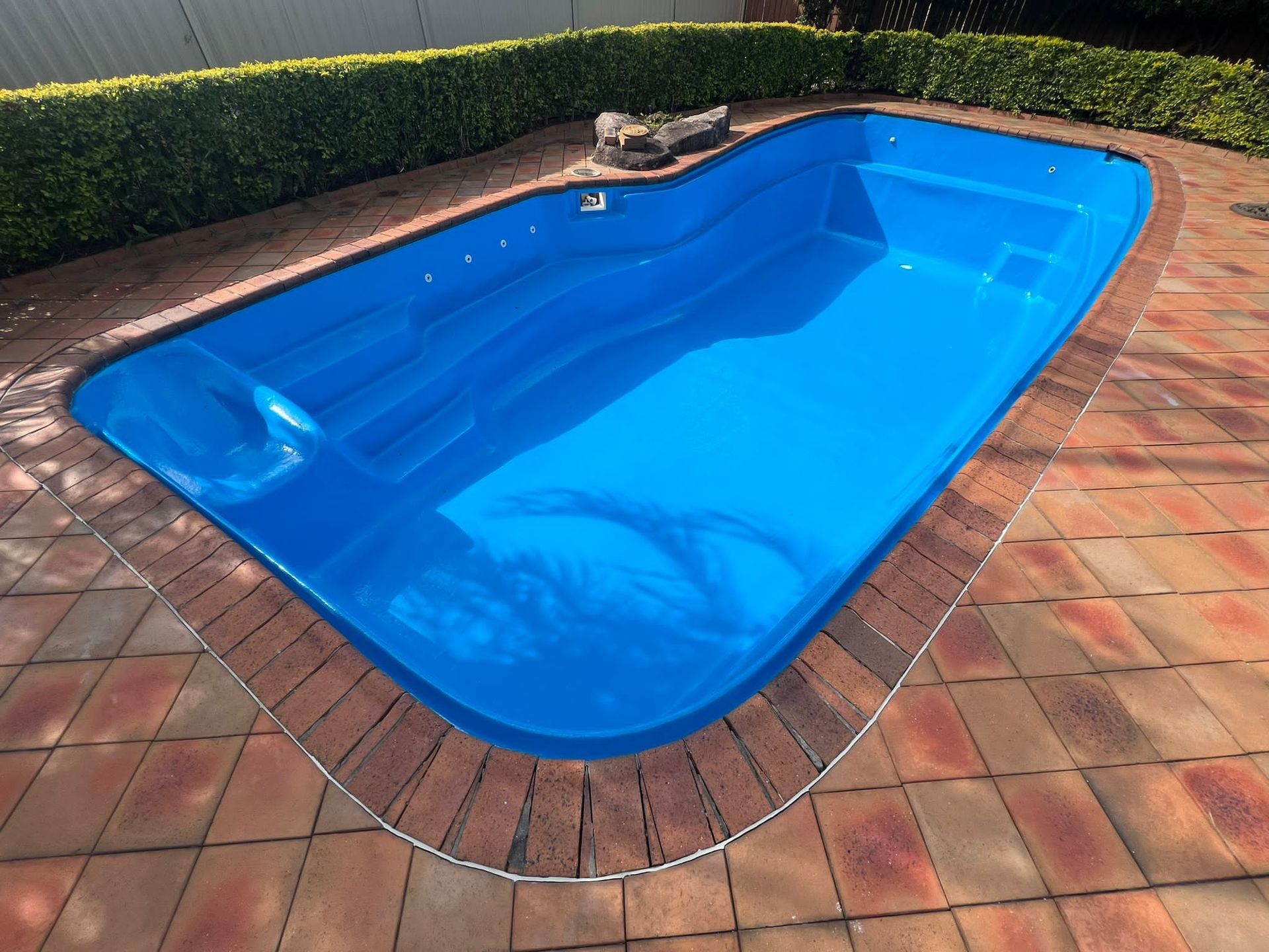 Empty blue swimming pool with a shallow ledge in bright sunlight, surrounded by tan deck and potted plants