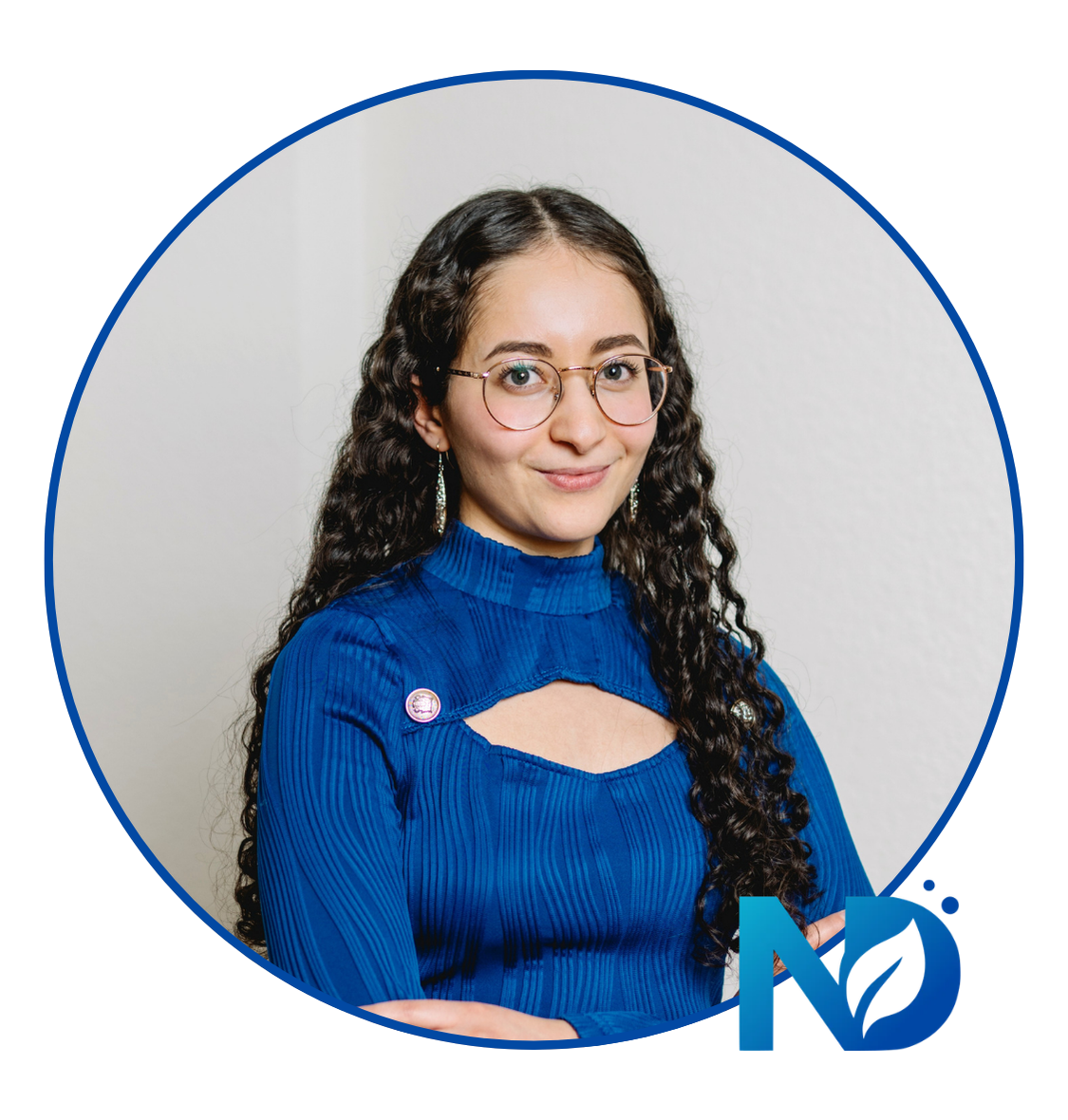 Femme à lunettes vêtue d'un haut bleu, souriante sur un fond gris clair, avec un petit logo bleu dans le coin