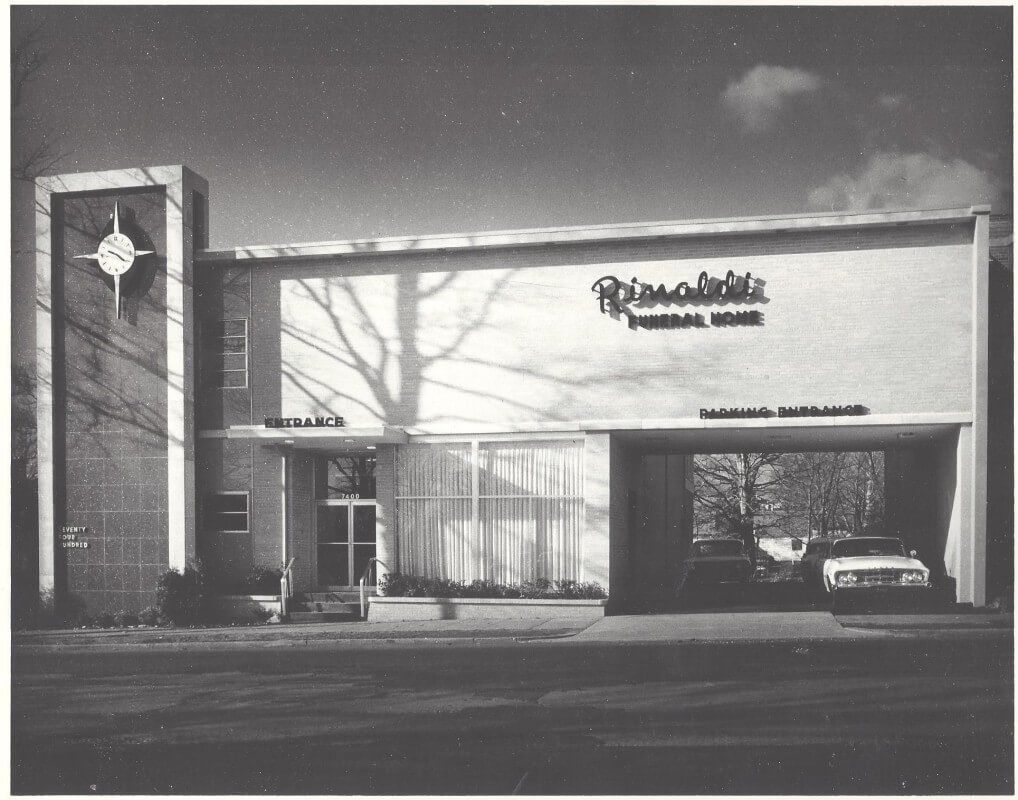 Black-and-white photo of a modern restaurant building with signage and a car parked under the entrance.
