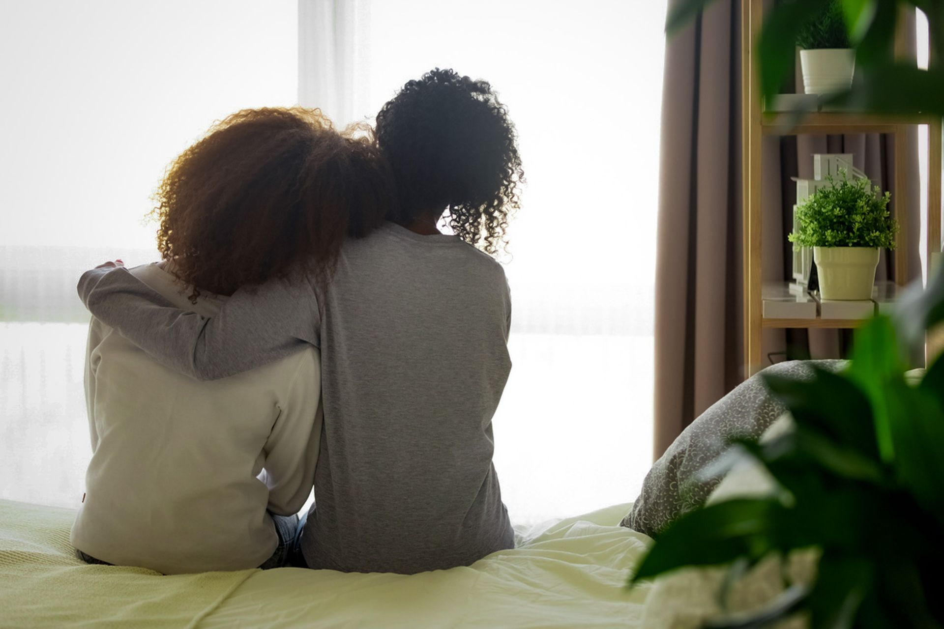 Two people sit side by side on a bed, looking out a bright window in a cozy room.