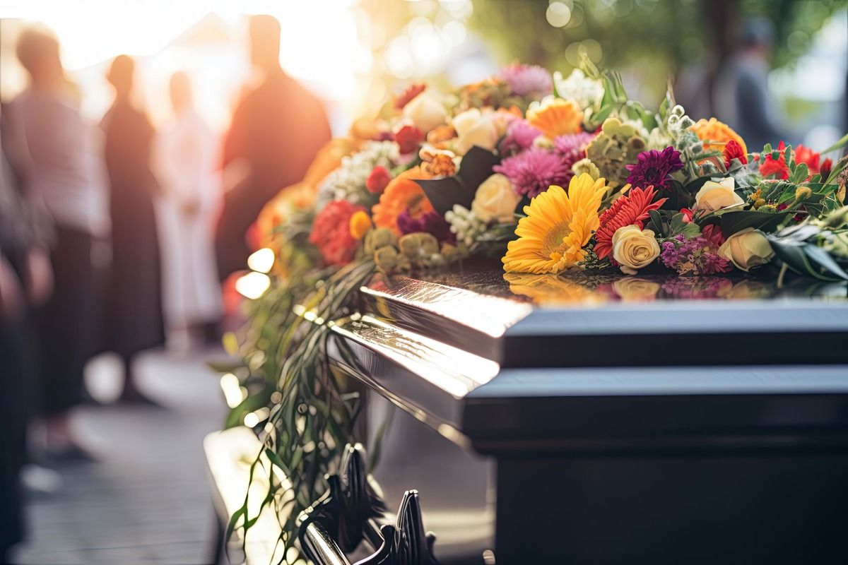 Flowers laid on a polished coffin at a graveside funeral, with mourners blurred in the background.