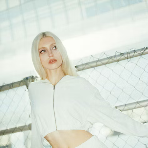 Blonde person in a white hoodie stands by a chain-link fence in bright daylight