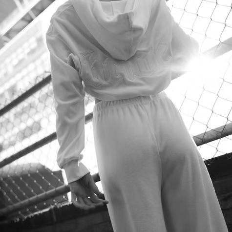 Person in a white outfit standing by a chain-link fence in black and white.