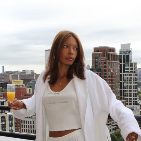 Woman in white on a rooftop holding a drink, with a city skyline in the background.