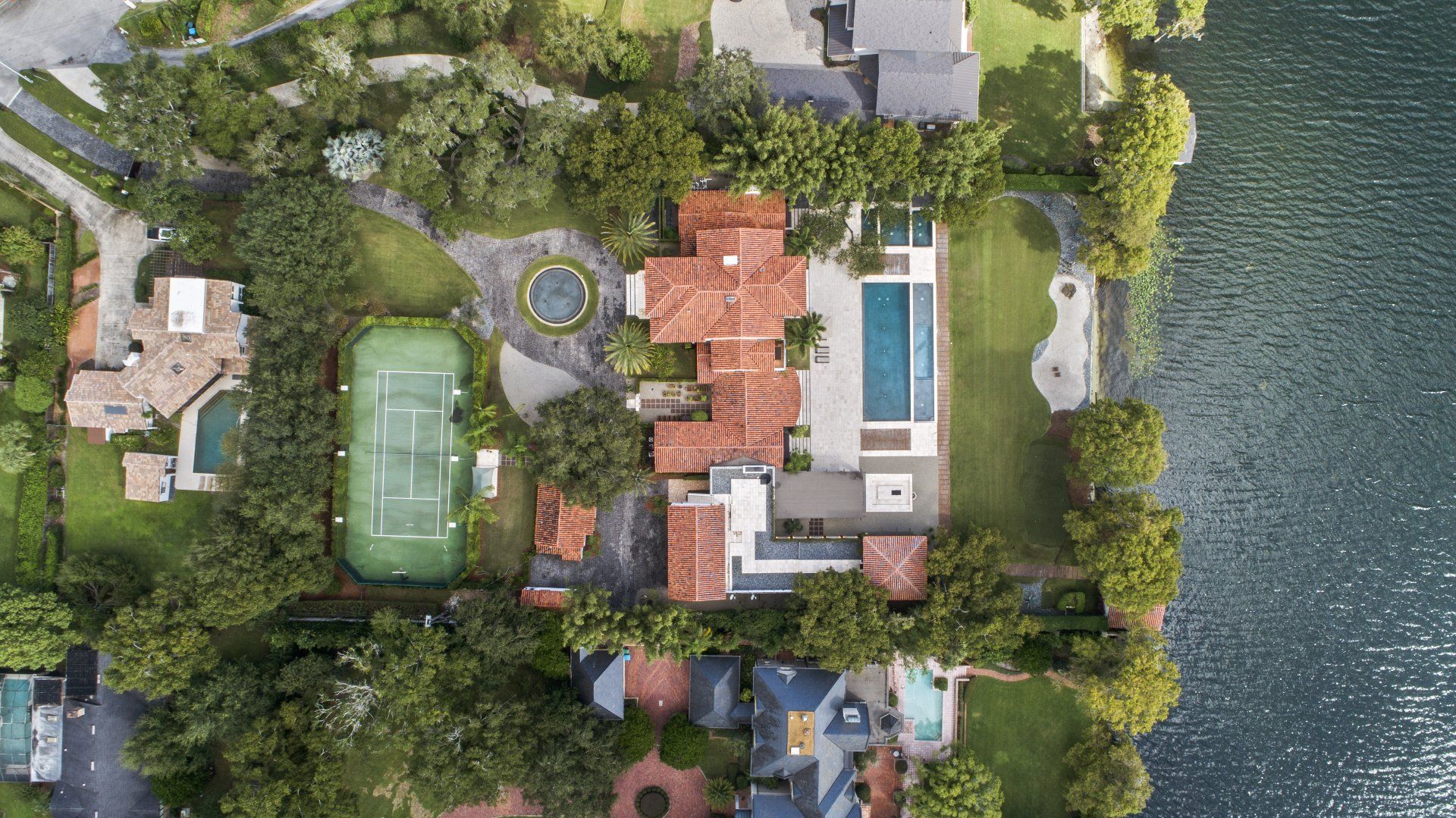 An aerial view of a house with a tennis court and a pool surrounded by trees.