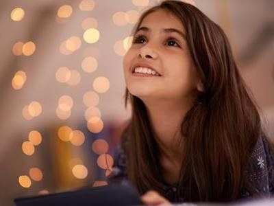 A smiling person looks upward while holding a tablet in front of a background of warm, out-of-focus golden fairy lights.
