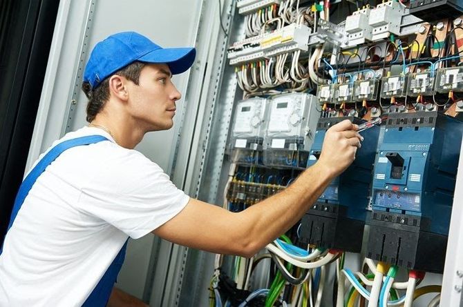 Technician in blue cap inspecting electrical control panel with tools and wiring.
