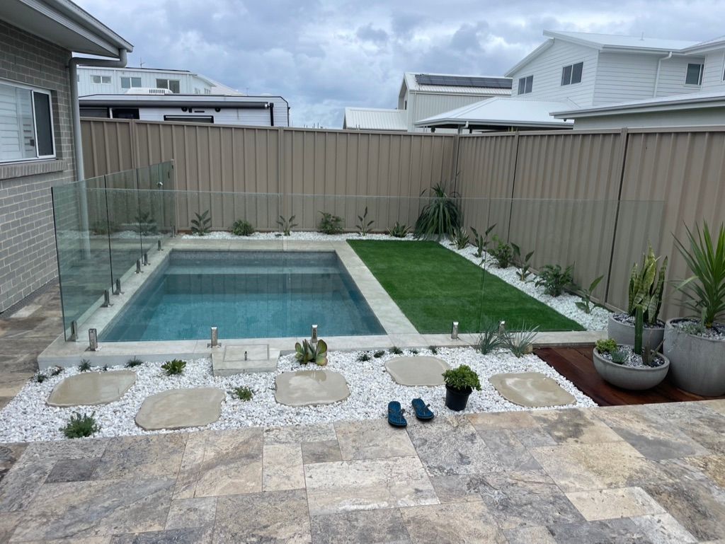 Backyard with pool, stone patio, white pebbles, grass, and plants. Beige fence and cloudy sky. — All Stars Plumbing In Dapto, NSW