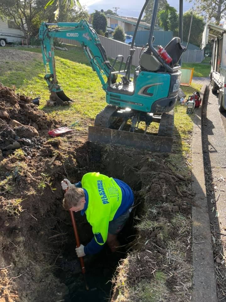 A person in a high-vis vest uses a shovel in a trench next to a small excavator on grass and a road. — All Stars Plumbing In Barrack Heights, NSW
