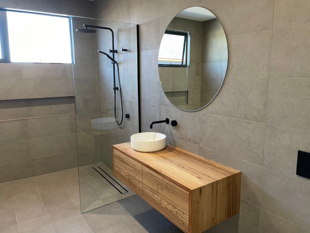 Modern bathroom with wooden vanity, round mirror, and glass shower enclosure. — All Stars Plumbing In Dapto, NSW