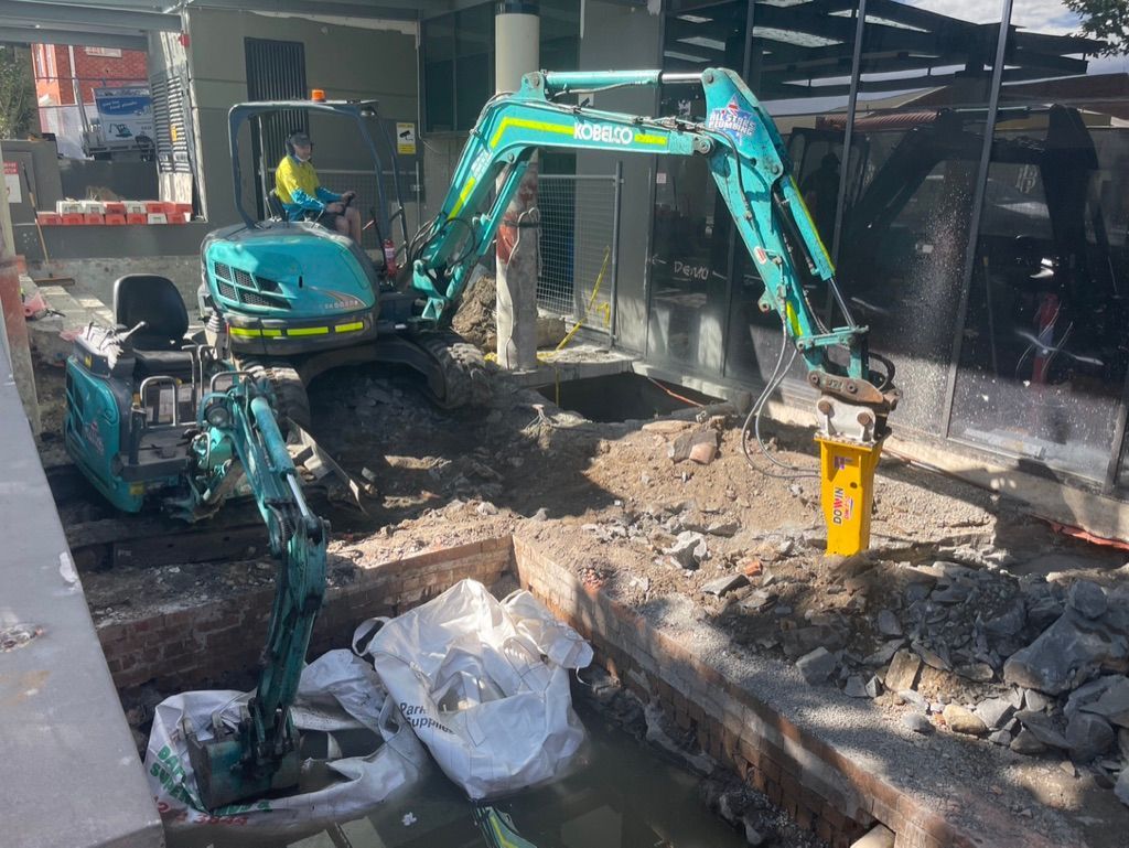 A small aqua excavator works in a construction site, next to a building. The excavator uses a jackhammer. — All Stars Plumbing In Dapto, NSW