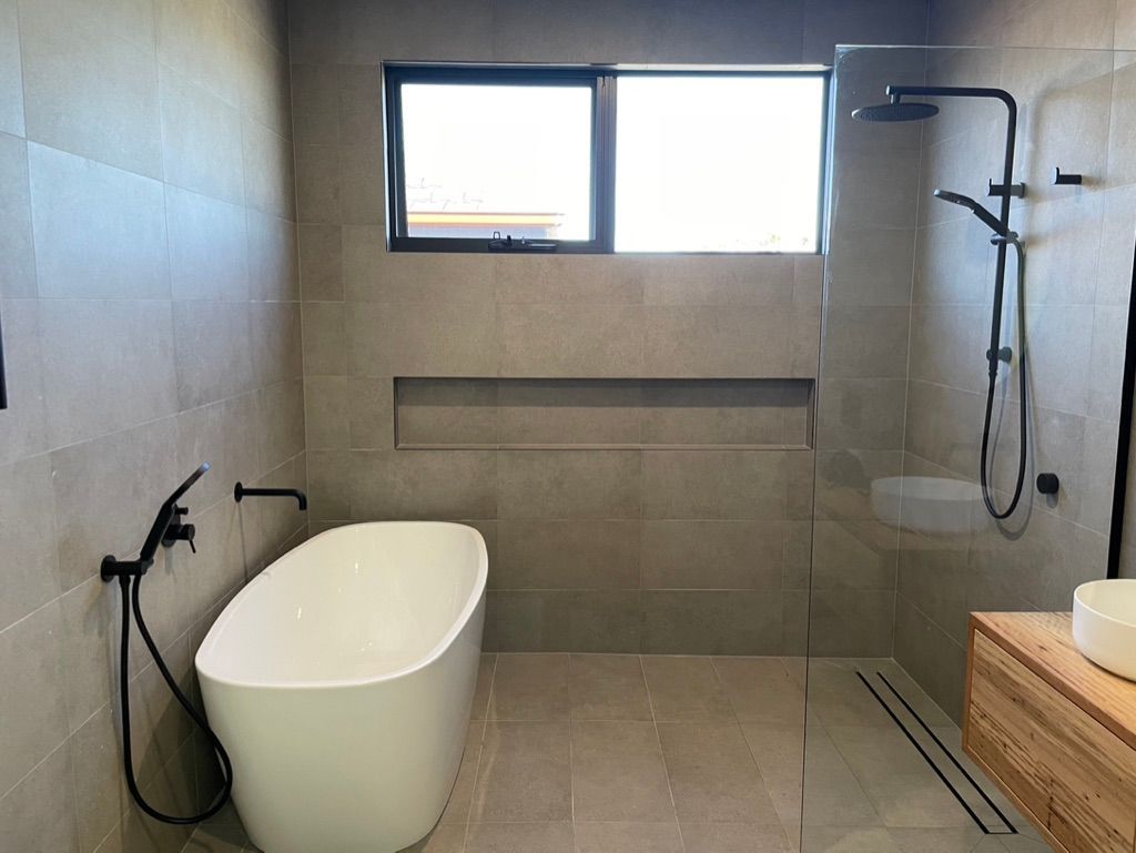 Modern bathroom with freestanding tub, shower, window, and wood vanity. Gray tiled walls and black fixtures. — All Stars Plumbing In Dapto, NSW