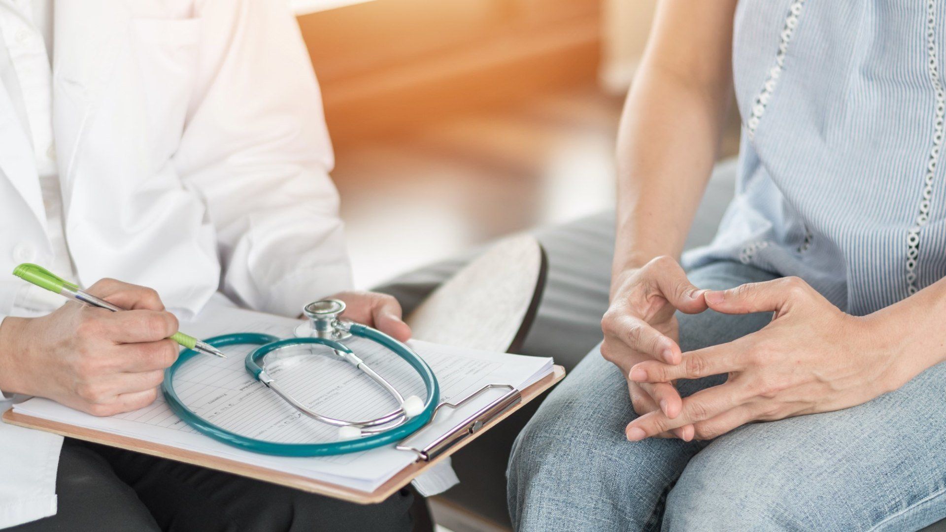 Doctor taking notes during a consultation with a seated patient, clipboard and stethoscope visible