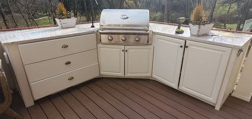 White outdoor kitchen island with grill, storage cabinets, and drawers on a wooden deck with potted plants.