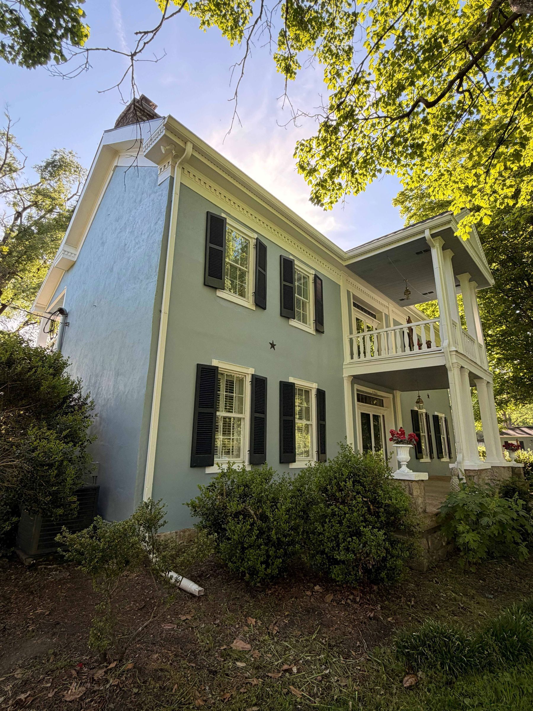 Light green two-story house with dark shutters and white porch, surrounded by trees and bushes