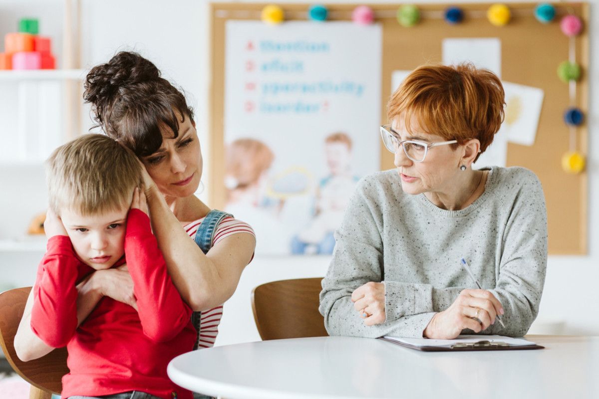 A woman comforts a child covering ears, while another woman with glasses sits at a table.