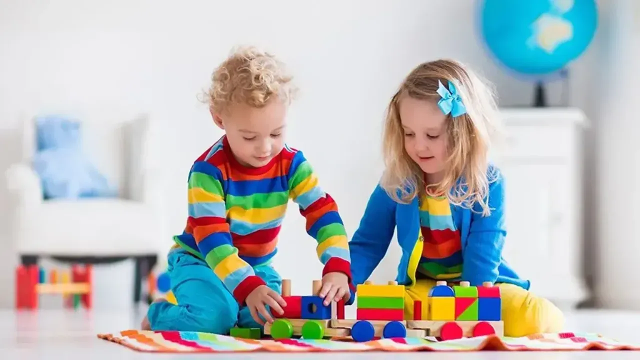 Two children playing with colorful wooden train and blocks on a rug.