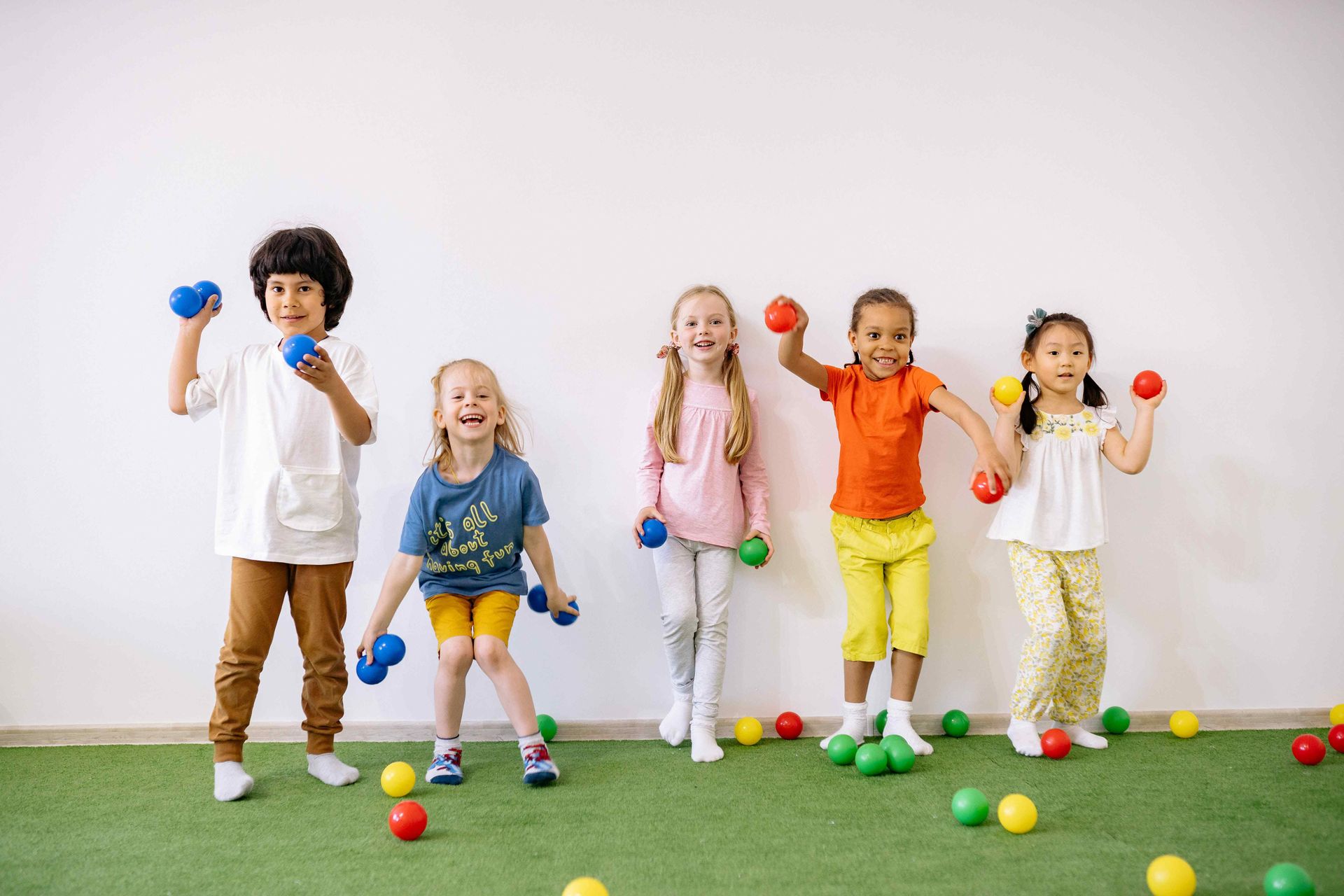 Five children holding colorful balls, smiling in front of a white wall, standing on green turf.