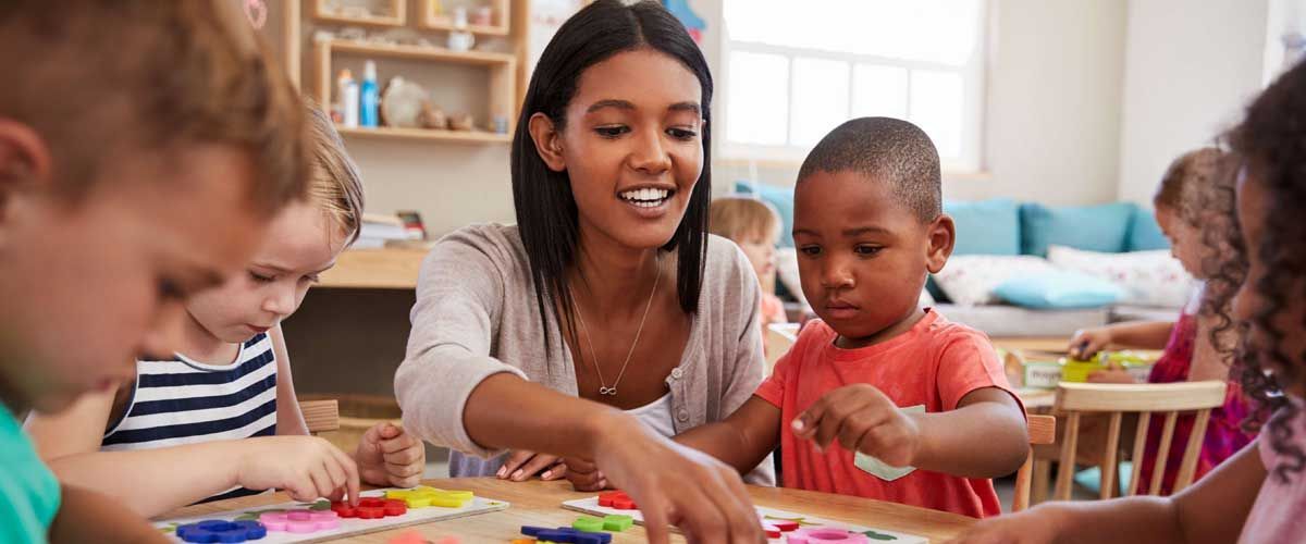 A teacher works with young children at a table, pointing at colorful shapes.