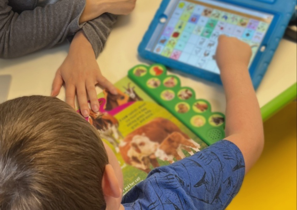 Child using a tablet with a communication app and a book on a table, another person's arm visible.