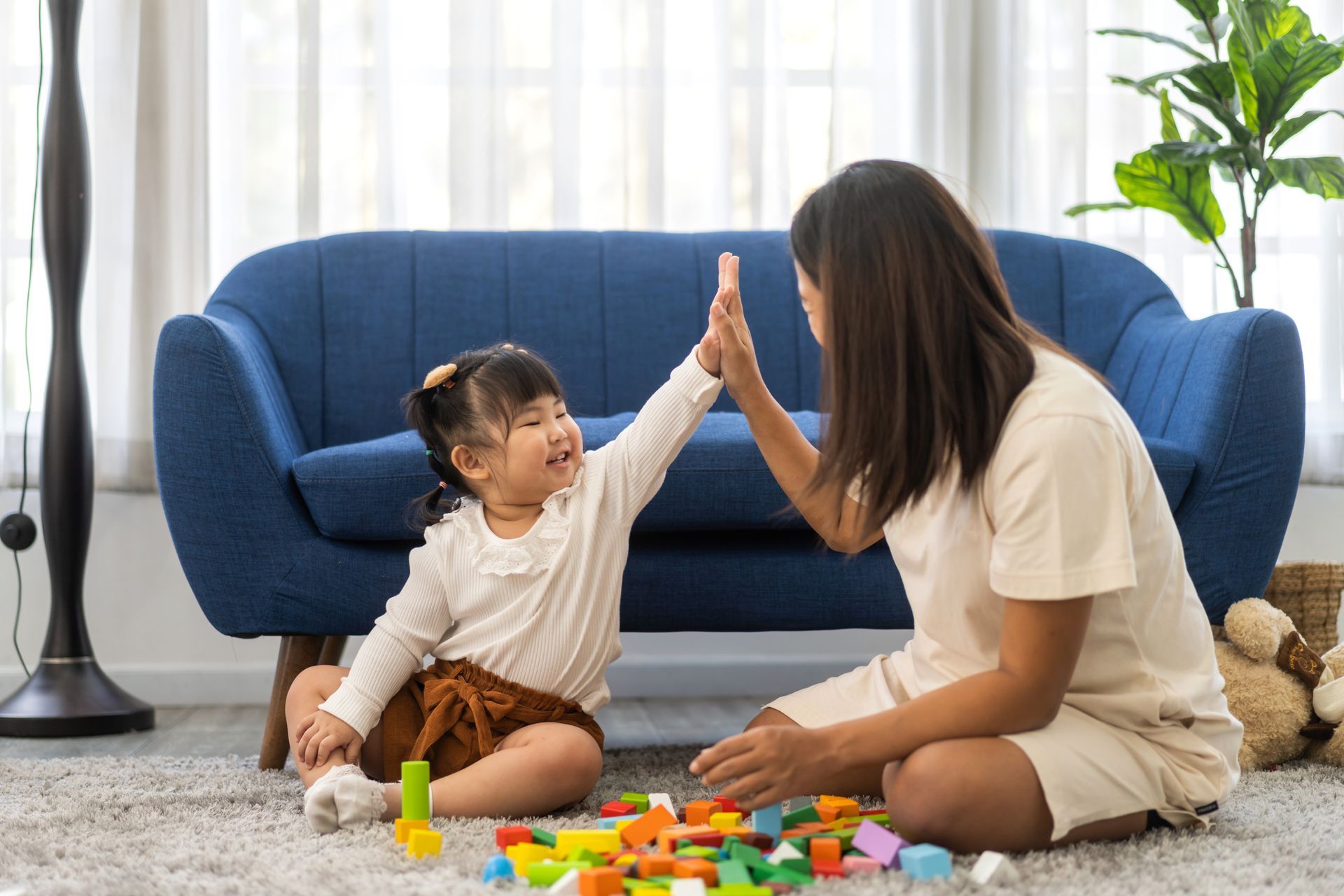 Woman and child high-five while playing with colorful blocks on the floor. Blue sofa in the background.