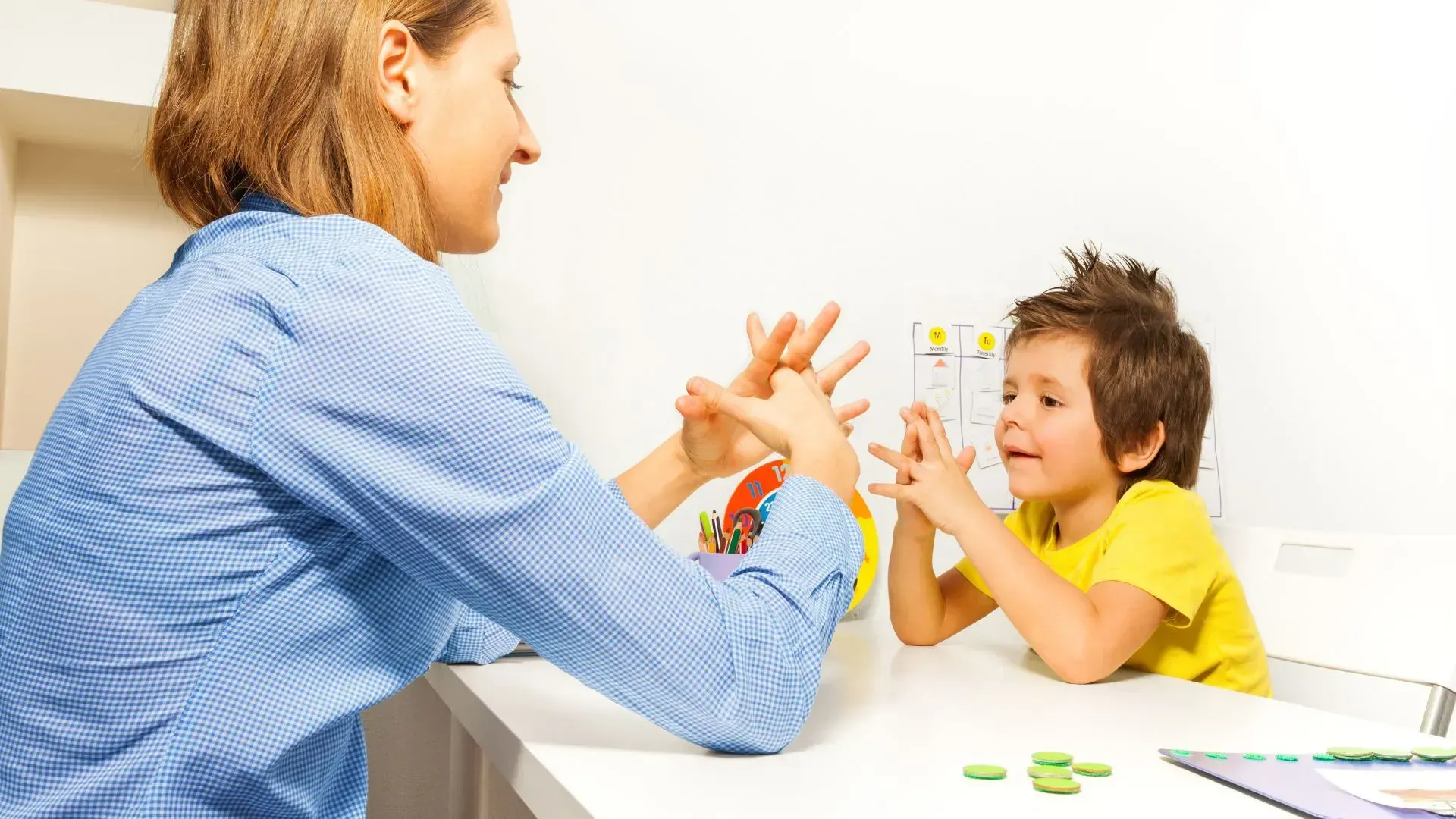 Woman and child performing a hand gesture exercise at a table, both smiling. White background.