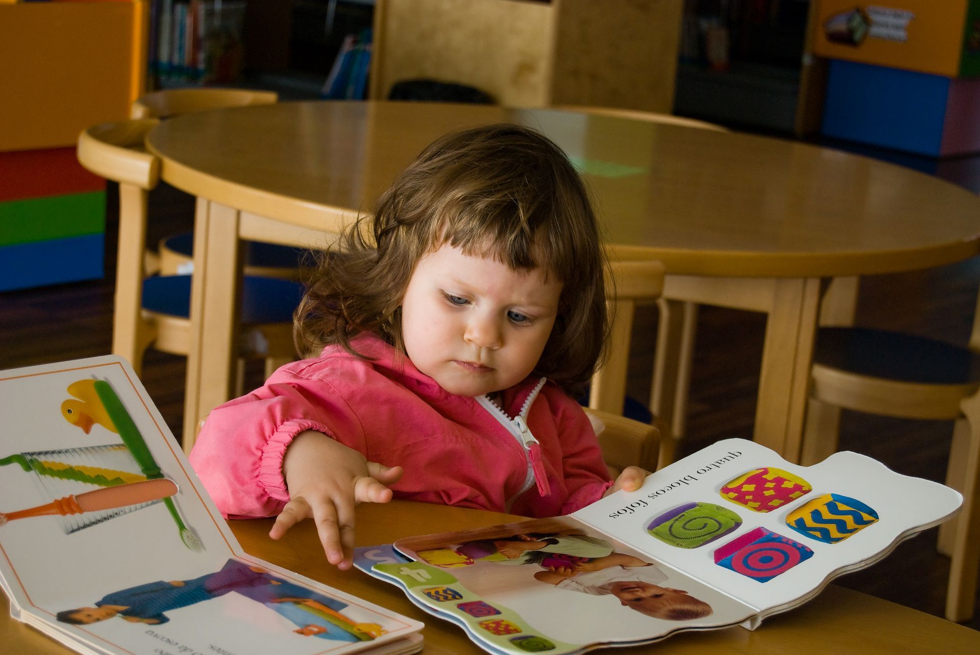 Young child in pink jacket reads colorful book at wooden table in a library.