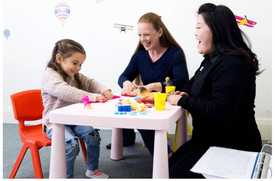 Child and two adults laugh, playing with toys at a small table in a brightly decorated room.