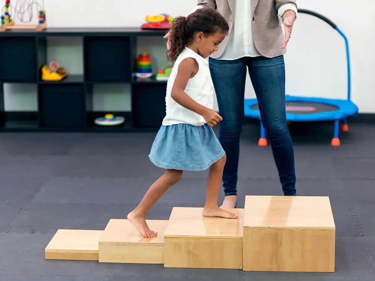 Girl walking up wooden steps with an adult's support in a therapy room.