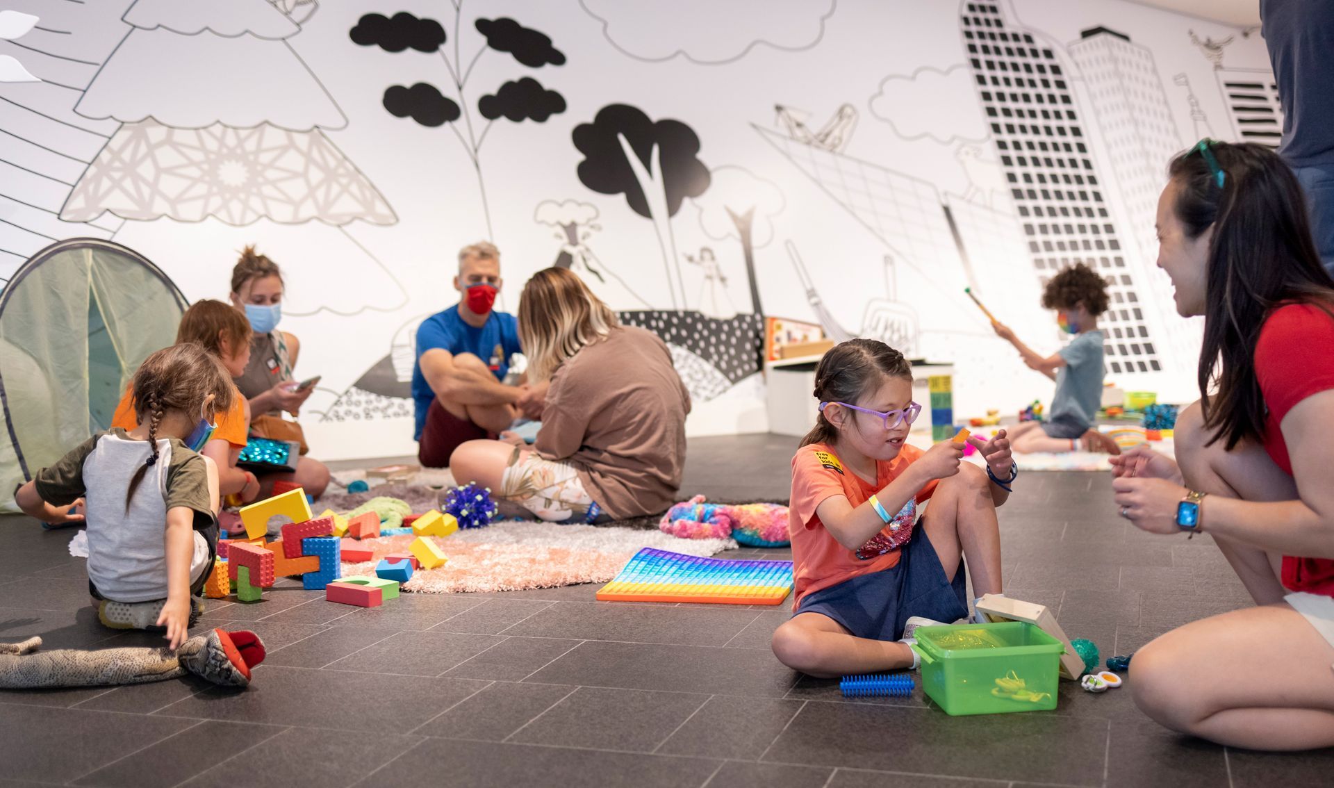 Children and adults playing on floor with toys in front of a mural. Some wear masks.