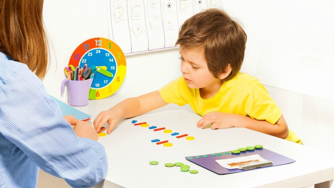 Boy in yellow shirt playing with colorful shapes at a table with an adult; clock and pencils in background.