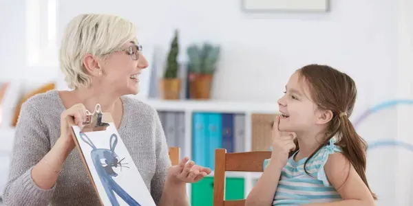Woman holding a clipboard with a drawing, talking to a child sitting at a table.