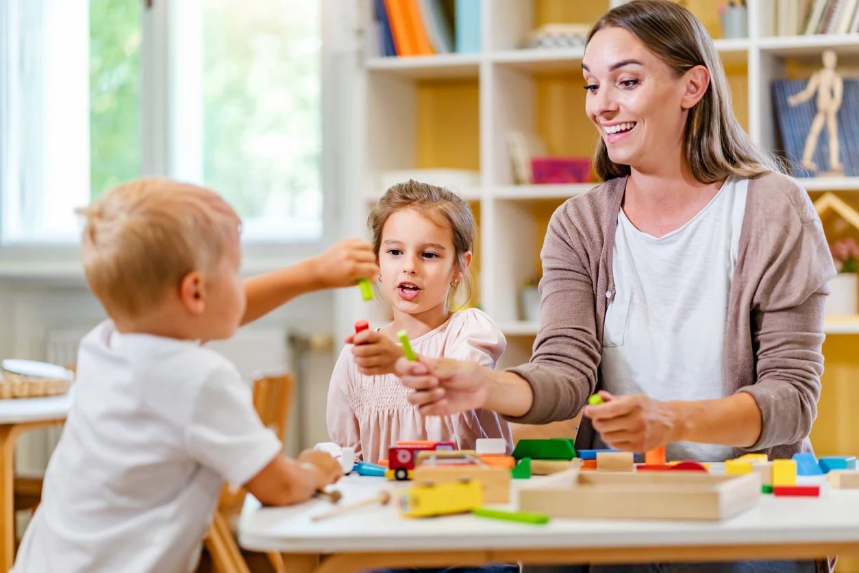 Teacher and two children playing with colorful blocks at a table. Bright room, natural light.