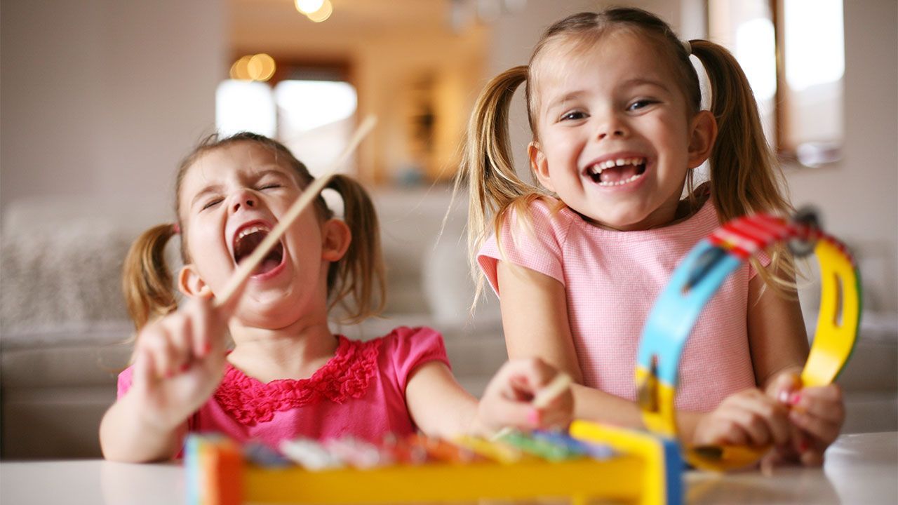 Two girls with pigtails playing musical instruments; one with a xylophone, the other with a tambourine, both laughing.