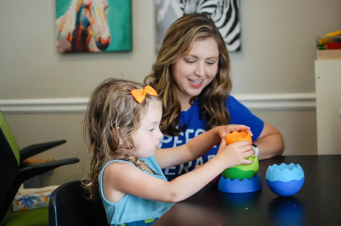 Child and woman playing with a stacking toy. Child wears orange bow; woman smiles.