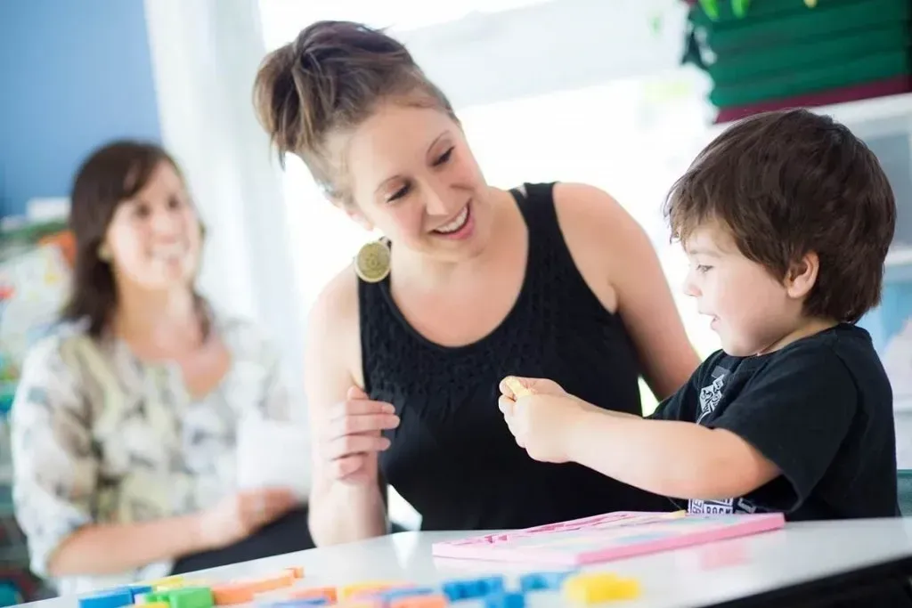 Woman smiles at child, assisting with a craft at a table, another woman watches in the background.