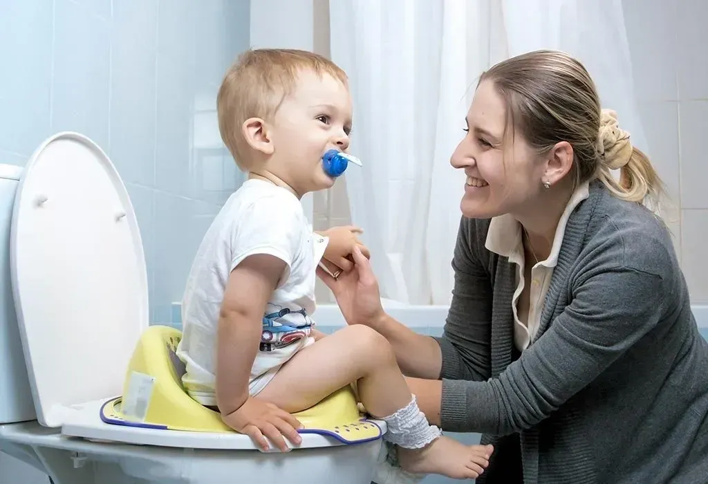 Woman helping a child sit on a child's toilet seat adapter on a toilet; child has a pacifier.